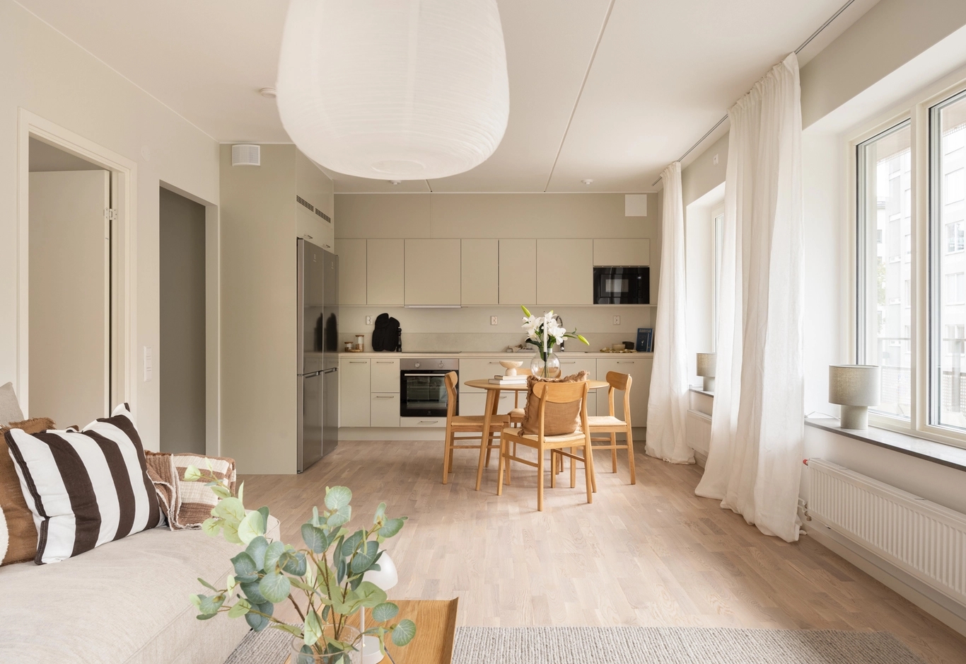 Modernt vardagsrum och kök i öppen planlösning med ljusa trägolv, neutrala möbler och stora fönster som släpper in naturligt ljus.