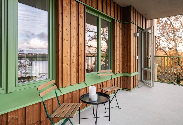 Träpanelad balkong med två stolar, ett litet runt bord, grönmålade fönster och utsikt över träd.