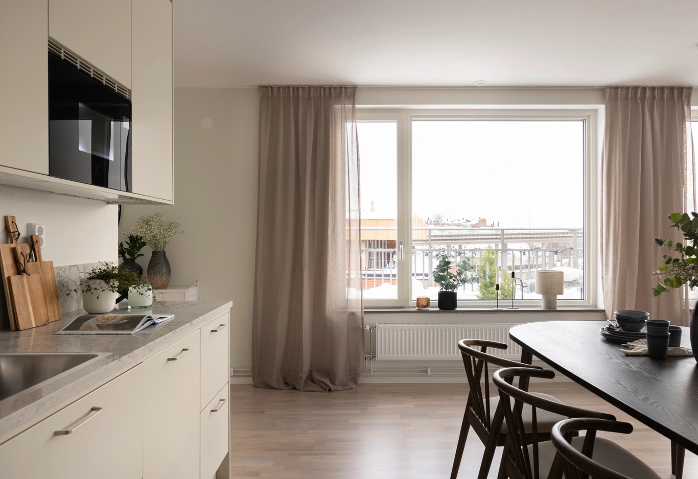 Modernt kök och matplats med trästolar, stort fönster och gardiner i neutrala färger.